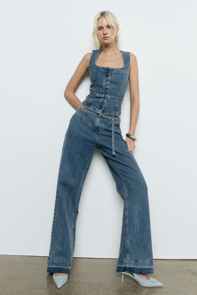 Model wears the SOVERE Dawn Denim Pant in Vintage Blue. Full-length mid-rise jeans with belt and contrast hem detail.